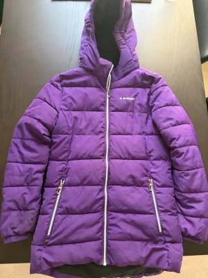 Lila dunjacka från Everest med huva - Snygg lila dunjacka från Everest med huva och två dragkedjeförsedda fickor framtill. Jackan har quiltad design, långa ärmar och dragkedja hela vägen upp. Perfekt för kalla vinterdagar och riktigt varm.