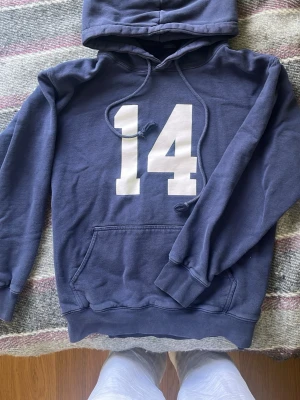 Mörkblå hoodie 14 - Mörkblå hoodie med stor vit siffra 14 på bröstet. Klassisk känguruficka framtill, huva med dragsko och ribbade muddar vid ärmar och nederkant. Perfekt för en avslappnad och sportig stil. Strl S-M, fråga längd så kan jag fixa 🤝