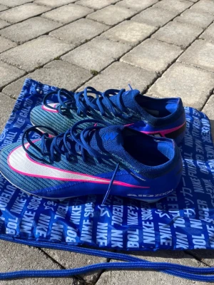 Blå Nike Mercurial fotbollsskor - Snygga blå Nike Mercurial fotbollsskor med rosa och vita detaljer. Skorna har en strömlinjeformad design, lågt skaft och snörning för bra passform. Tillverkade i syntetmaterial för lätt vikt och bra bollkontroll. Perfekta för dig som vill sticka ut på planen.