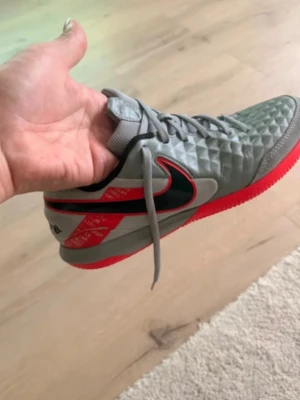 Gråa och röda Nike fotbollsskor - Säljer ett par gråa Nike fotbollsskor med svart swoosh och röda detaljer. Skorna har en mönstrad ovandel och röd sula som ger bra grepp på planen. Snygg design med snörning och hög komfort för dig som vill sticka ut på fotbollsplanen.