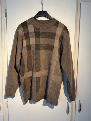 Biegie stickad tröja med rutmönster - Snygg stickad tröja i bruna och beige toner med ett stort rutmönster framtill. Tröjan har rund halsringning, långa ärmar och ribbade muddar vid ärmslut och nederkant. Perfekt för dig som gillar en stilren och trendig look.