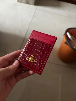 Röd kortfodral med krokopräglat mönster - Snyggt rött kortfodral i konstläder med krokopräglat mönster på framsidan och guldfärgad metallogo. Fodralet har plats för flera kort och en slät baksida. Perfekt för dig som vill ha en stilren och trendig accessoar i fickan.