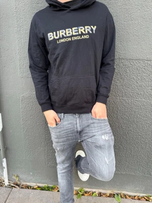 Svart Burberry hoodie med tryck - Svart hoodie från Burberry med stort logotryck i gult och vitt på bröstet. Klassisk huva och känguruficka framtill. Mjuk bomullskänsla och avslappnad passform, perfekt för en stilren och trendig look.