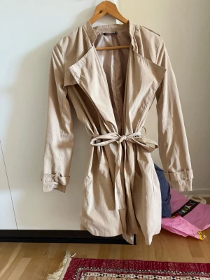 Beige trenchcoat med knytskärp - Stilren beige trenchcoat med klassisk krage, knytskärp i midjan och dekorativa knappar på ärmarna. Lätt material och avslappnad passform, perfekt för lager-på-lager. Snygg detalj med breda slag framtill. Ger riktigt bra passform! Storlek 36 / S 