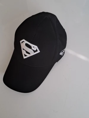 Svart keps med Superman-logga - Snygg svart keps med vit broderad Superman-logga framtill och på sidan. Klassisk böjd skärm och justerbar passform. Tillverkad i slitstarkt tyg som passar perfekt till streetwear eller casual outfits.