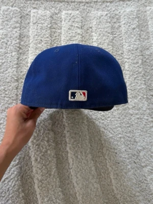 Mörkblå MLB fitted-keps med broderad logga - Säljer en mörkblå keps med broderad MLB-logga bak och vit knapp på toppen. Klassisk basebollmodell med böjd skärm och ventilerande hål. Kepsen är gjord i ett tåligt tygmaterial och har en stilren look.