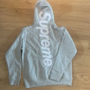 Grå Supreme hoodie  - Säljer en grå hoodie från Supreme med stor vit logga längs ryggen. Klassisk känguruficka framtill, dragsko i huvan och ribbade muddar. Perfekt för dig som gillar streetwear och vill sticka ut med en clean men ändå fet design.