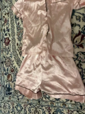 Rosa satinpyjamas med korta ärmar - Söt pyjamas i glansigt rosa satinmaterial med kortärmad skjorta och matchande shorts. Svarta passpoaldetaljer och diskreta hjärtmönster ger en gullig touch. Perfekt för mysiga kvällar hemma.