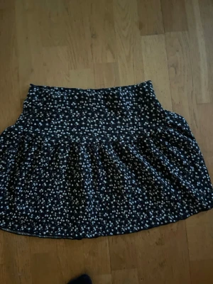 Svart blommig kjol från Gina Tricot - Söt svart kortkjol från Gina Tricot med små vita blommor. Kjolen har en bred resår i midjan och ett lätt utställt fall. Perfekt för dig som gillar en avslappnad men ändå trendig stil.