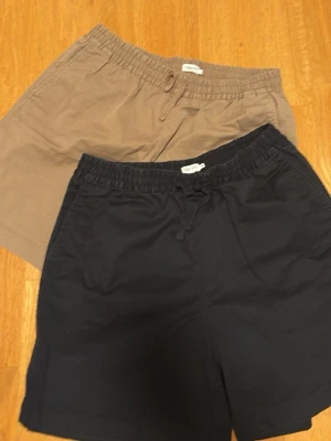 Två par shorts med resårmidja - Säljer två par shorts, ett par i beige och ett par i mörkblått. Båda har resårmidja med snörning och sidofickor. Perfekta för sommaren och tillverkade i mjuk bomull för maximal komfort.
