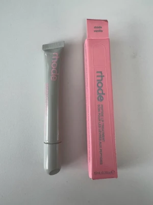 Rhode Peptide Lip Treatment Vanilla - Rhode Peptide Lip Treatment i doften Vanilla. Läppbalsamet kommer i en stilren grå tub med rosa text och matchande rosa förpackning. Tuben rymmer 10 ml och har en smidig applikator. Perfekt för att återfukta och vårda läpparna.