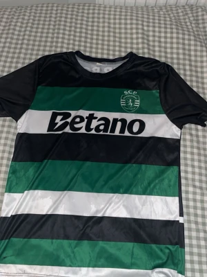 Sporting CP fotbollströja svart/grön - Säljer en officiell Sporting CP fotbollströja med breda svarta, gröna och vita ränder. Tryck med Betano på framsidan och Gyökeres samt nummer 9 på ryggen. Tröjan har korta ärmar och är tillverkad i ett glansigt, snabbtorkande material.