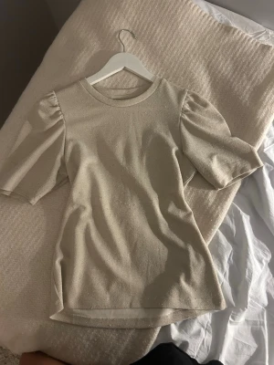 Guldig topp med puffärm och glitter - Trendig guldig topp med puffärmar och glittereffekt. Rund halsringning och rak passform ger en stilren look. Perfekt för dig som vill ha något extra till din outfit. Materialet känns mjukt och har en lätt glans. Den kan se lite nopprig ut på magen men inget som jag tror att man tänker på!💓