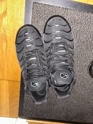Svarta Nike tns - Snygga svarta sneakers med silvriga detaljer och vågformade mönster på ovandelen. Skorna har en sportig look med synlig dämpning i sulan och svarta skosnören. Materialet är en mix av textil och syntet, perfekt för en streetwear-stil. Storlek 42 passar även 41