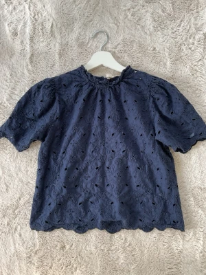 Mörkblå blus - Söt mörkblå blus med broderade hål och puffiga korta ärmar. Blusen har rundad hals med liten volangkant och vågig nederkant. Tillverkad i ett lätt tyg som ger en romantisk känsla.