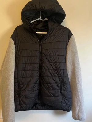 Svart cardigan med grå ärmar - Säljer en stilren svart pufferjacka med grå sweatshirt-ärmar och huva. Jackan har dragkedja framtill och två fickor på sidorna. Kroppen är quiltad och vadderad för extra värme, medan ärmarna är i mjukt sweatshirtmaterial. Perfekt för kalla dagar.