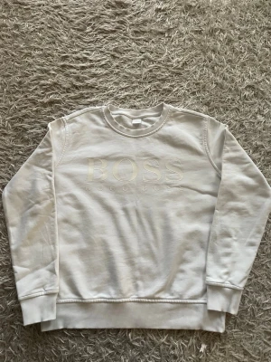 Vit sweatshirt från Hugo Boss - Säljer en stilren vit sweatshirt från Hugo Boss med broderad logga framtill. Tröjan har rund halsringning, långa ärmar och ribbade muddar vid ärmslut och nederkant. Tillverkad i mjukt material som känns skönt mot huden. Perfekt för dig som gillar klassisk och clean stil.