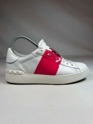 Valentino vita sneakers med rosa detalj - Snygga vita sneakers från Valentino med en bred, glansig rosa remsa över mitten och matchande rosa detaljer vid snörningen. Skorna har en chunky vit sula och nitar baktill. Tillverkade i läder för en lyxig känsla och stilren look. Priset är ej hugget i sten så kom med bud🤝