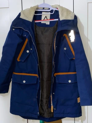 Mörkblå parkas med fuskpälsfoder - Snygg mörkblå parkas med varm fuskpäls i huvan och orange detaljer på fickorna. Jackan har dragkedja, tryckknappar och fyra stora fickor framtill. Perfekt för kalla dagar och har en längre modell som skyddar mot vind och kyla. Mått: längden är ca 88cm, axelbredden är ca 40cm och ärmlängden är ca 66cm 