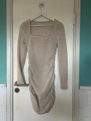 Randig beige och vit kortklänning - Kortklänning med beige och vita ränder, långärmad och med fyrkantig ringning. Klänningen har en åtsittande passform och rynkade detaljer på sidan för en snygg siluett. Perfekt för dig som gillar en enkel men trendig stil.