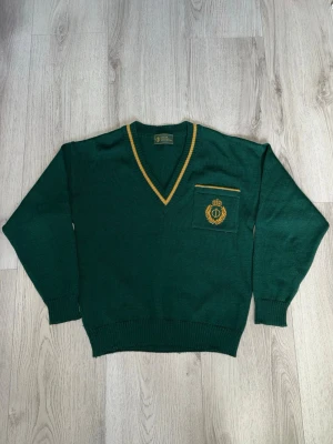 Grön stickad tröja Oscar Jacobson - Klassisk mörkgrön V-neck-tröja från Oscar Jacobson med guldfärgade detaljer i kragen och en fin broderad logga/sköld på bröstet. Ger en riktigt snygg "preppy" och collegestil! 