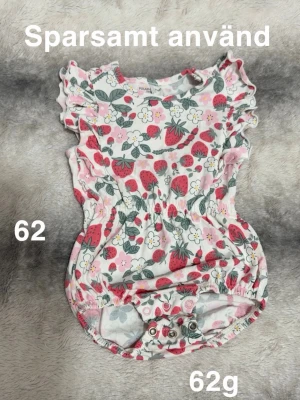 Söt jordgubbsbody med volanger - Supersöt body med volangärmar och tryck av jordgubbar och blommor i rosa, rött, grönt och vitt. Smidig knäppning i grenen och resår i midjan för skön passform. Perfekt för dig som gillar färgglada och lekfulla plagg till de minsta.🌸 Sparsamt använd. Storlek 62 