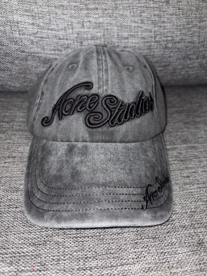 Grå keps från Acne Studios - Snygg grå keps från Acne Studios med broderad logga framtill och på skärmen. Kepsen har en klassisk böjd skärm och justerbar rem bak. Tillverkad i mjukt bomullstyg med en tvättad look som ger en avslappnad vibe.