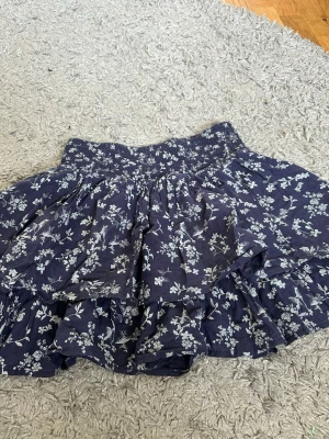 Blommig volangkjol från H&M L.O.G.G. - Mörkblå kortkjol från H&M L.O.G.G. med vitt blommönster och volangdetaljer. Kjolen har bred resår i midjan och är gjord i mjuk bomull, perfekt för en avslappnad och trendig look.