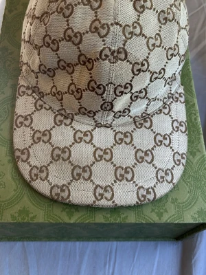 Äkta Beige GG-mönstrad keps från Gucci - Snygg beige keps från Gucci med det klassiska GG-monogrammet i brunt över hela kepsen. Tillverkad i canvasmaterial med mjukt foder på insidan och böjd skärm. Perfekt för dig som vill ha en exklusiv och trendig accessoar.