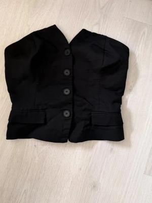 Svart croppad väst med knappar - Snygg svart croppad väst med fyra stora knappar framtill och dekorativa fickor. Västen har en figurnära passform och smockdetalj i ryggen för extra komfort. Perfekt att styla över en skjorta eller t-shirt för en trendig look.