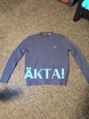 Buda på en äkta Blå kabelstickad tröja Polo Ralph Lauren - Snygg blå kabelstickad tröja från Polo Ralph Lauren med den klassiska gula loggan broderad på bröstet. Tröjan har rund halsringning, långa ärmar och ribbade muddar. Perfekt för lager på lager och en stilren look.