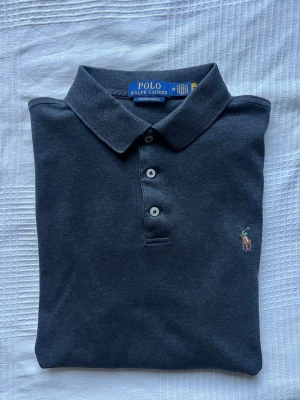 Mörkgrå pikétröja från Polo Ralph Lauren - Klassisk mörkgrå pikétröja från Polo Ralph Lauren i medium med modellen Custom Slim Fit. Tröjan har krage, tre knappar framtill och den ikoniska broderade loggan på bröstet. Perfekt för en stilren och avslappnad look.