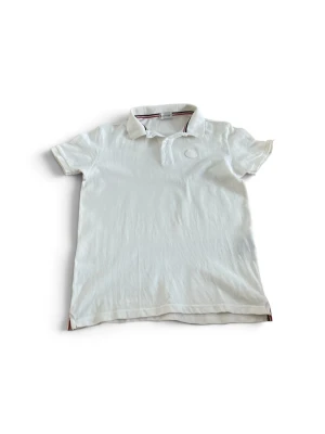 Vit pikétröja från Moncler - Klassisk vit pikétröja från Moncler med krage och knappslå. Tröjan har Moncler-logga ton-i-ton på bröstet och diskreta ränder i kragen. Perfekt till en clean och stilren look.