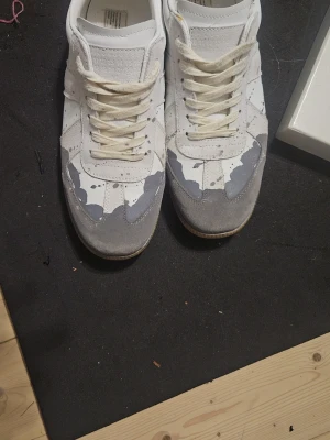 Vita sneakers med grå detaljer - Säljer ett par vita sneakers med grå mockadetaljer och unika färgstänk i grått och svart. Skorna har vita snören och en klassisk låg siluett med gummisula. Perfekta för dig som vill sticka ut med en kreativ och trendig look.