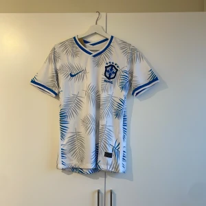 Vit och blå fotbollströja från Nike - Snygg vit fotbollströja från Nike med blå palmbladsmönster och detaljer. Tröjan har korta ärmar och en rund halsringning. Perfekt för fotbollsfans som vill ha en unik stil på planen. Brasilien 2022 special edition