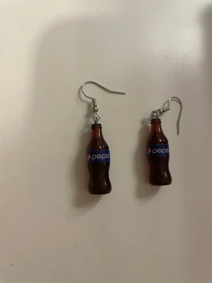 Örhängen med Pepsi-flaskor - Unika örhängen formade som små Pepsi-flaskor i brunt och blått. Tillverkade i plast med metallkrokar. Perfekt accessoar för dig som gillar roliga och annorlunda smycken.