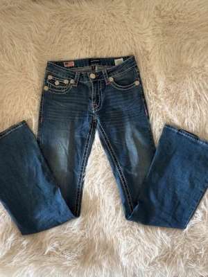 Blå bootcut jeans med kontrastsömmar - Säljer ett par blå bootcut jeans från True Religion med vita kontrastsömmar och detaljerade bakfickor med knappar. Jeansen har klassisk femficksdesign och är tillverkade i denim med lätt tvättad look. Perfekta för en avslappnad och trendig stil.