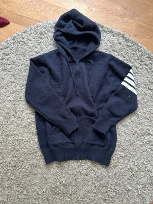 Mörkblå hoodie med segelbåt - Mörkblå stickad hoodie med dragkedja och huva. Ena ärmen har fyra vita ränder och på ryggen finns ett stort segelbåtsmotiv i vitt med en röd detalj. Hoodien har fickor framtill och ribbade muddar. Perfekt för en avslappnad och nautisk stil.