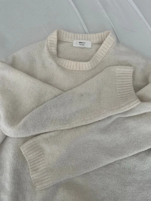 Beige stickad tröja från Gina - Mysig beige stickad tröja med rund halsringning och ribbade muddar vid ärmslut och hals. Tröjan har en enkel och stilren design, perfekt för lager-på-lager och passar till det mesta.