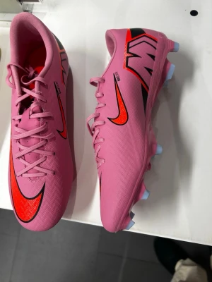 Rosa Nike fotbollsskor med dobbar - Säljer ett par snygga rosa fotbollsskor från Nike med röda och svarta detaljer. Skorna har klassisk snörning, låg profil och dobbar för bra grepp på planen. Materialet är syntetiskt och designen är sportig och modern.