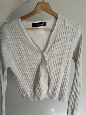 Vit ribbad kofta  - Snygg vit ribbad kofta med v-ringning och två knappar framtill. Modellen är croppad och har långa ärmar samt breda muddar. Perfekt att styla över ett linne eller t-shirt för en trendig look.