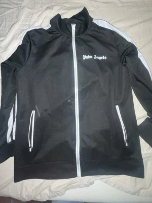 Palm Angels svart track jacket - Snygg svart track jacket från Palm Angels med vita detaljer och dragkedja framtill. Jackan har vita ränder längs ärmarna, ståkrage och två fickor med dragkedja. Perfekt för en sportig och trendig look.