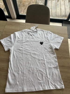 Cdg t-shirt - Säljer en vit t-shirt med korta ärmar och en svart broderad hjärtdetalj med ögon på bröstet. T-shirten har en klassisk passform och är gjord i mjuk bomull. Perfekt till jeans eller shorts för en clean och stilren look. Passar även S!