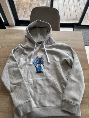 Grå hoodie med Polo Bear från Ralph Lauren - Snygg grå hoodie från Ralph Lauren med den ikoniska Polo Bear-figuren tryckt på bröstet. Hoodien har vit dragsko, känguruficka och ribbade muddar. Perfekt för en avslappnad och trendig stil.