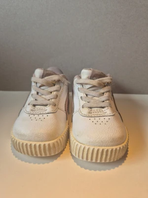 Vita sneakers med rosa detalj - Säljer ett par vita sneakers från Puma med ljusrosa sidostripes och beige detaljer. Skorna har klassisk låg modell, snörning och räfflad sula. Perfekta för en avslappnad och trendig stil.