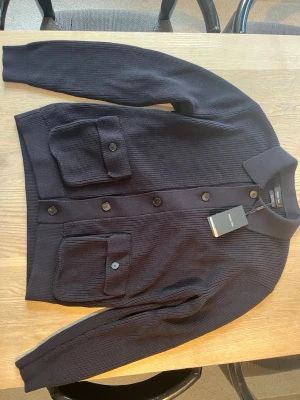 Mörkblå stickad cardigan med fickor från Massimo Dutti  - Snygg mörkblå cardiganjacka i stickad bomull från med två stora fickor framtill och knappar längs hela framsidan. Jackan har en klassisk krage och ribbade muddar vid ärmslut och nederkant. Perfekt för lager på lager och en stilren look. Tröjan är helt ny och helt oanvänd 