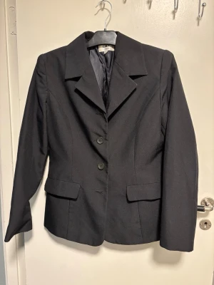 Svart kavaj från C.N.Y. Women - Stilren svart kavaj från C.N.Y. Women med klassisk krage, tre knappar framtill och två fickor med lock. Kavajen har figurnära passform och är helfodrad för extra komfort. Perfekt för en snygg och uppklädd look.