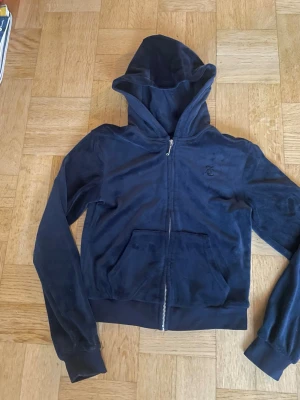 Mörkblå Juicy couture  - Mysig mörkblå hoodie i velour med dragkedja framtill och  huva. Tröjan har fickor på magen och en diskret broderad logga på bröstet. Perfekt för en avslappnad och trendig look.