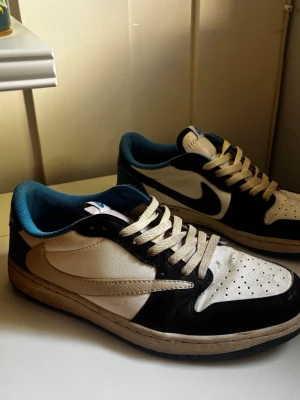 Svarta och vita sneakers med blå detaljer Travis scott  - Snygga sneakers i svart och vitt med blå insida och klassisk låg profil. Skorna har perforerad tå, vita skosnören och tydliga kontrastsömmar. Materialet är läderimitation och yttersulan är beige. Perfekta för en streetwear-look.
