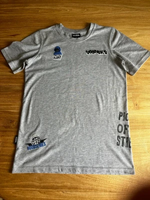 Grå t-shirt med tryck från Hoodrich - Snygg grå t-shirt från Hoodrich med flera tryck på bröstet och nedtill, bland annat texten 'HOODSERIES' och grafiska detaljer i svart och blått. T-shirten har rund hals och korta ärmar, perfekt för en avslappnad streetwear-look.